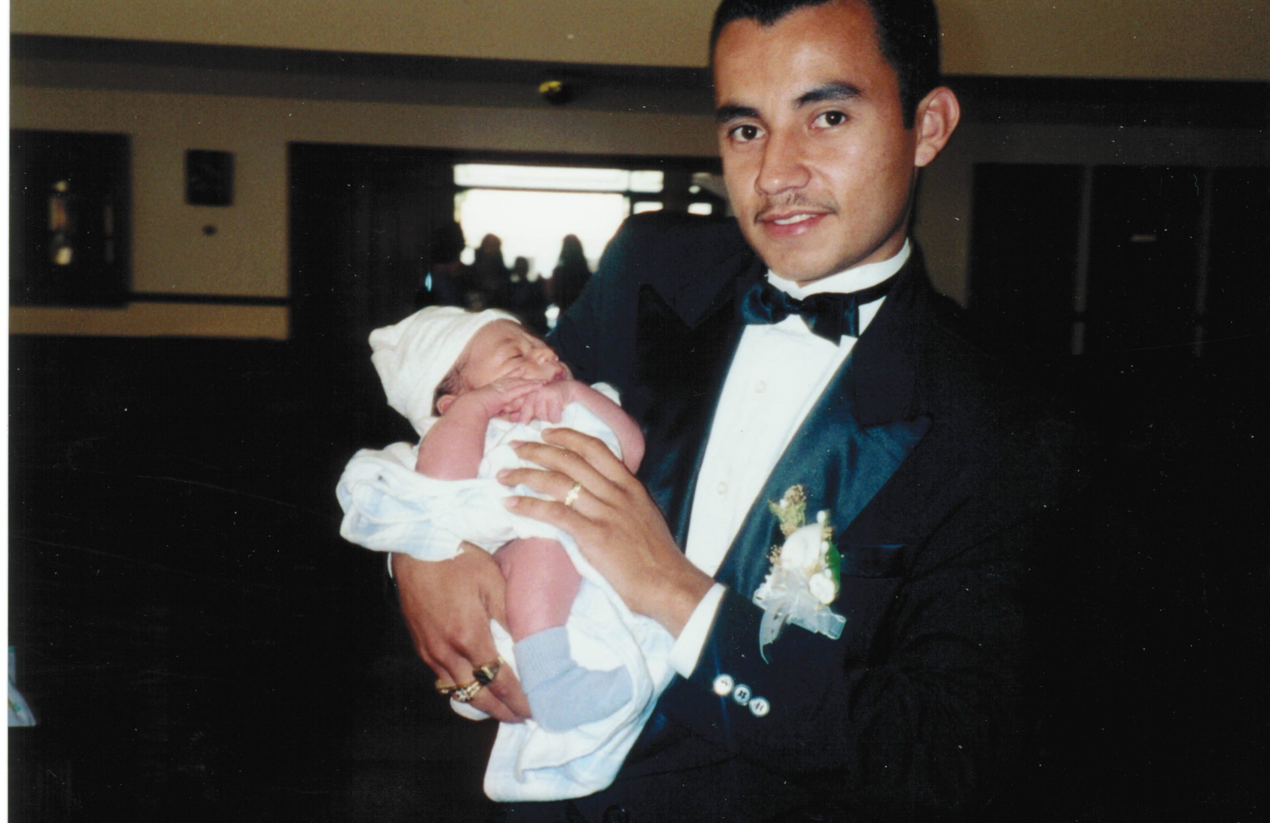 Areli Escobar, a Latinx man, poses with a baby while wearing a black tuxedo and bow-tie.