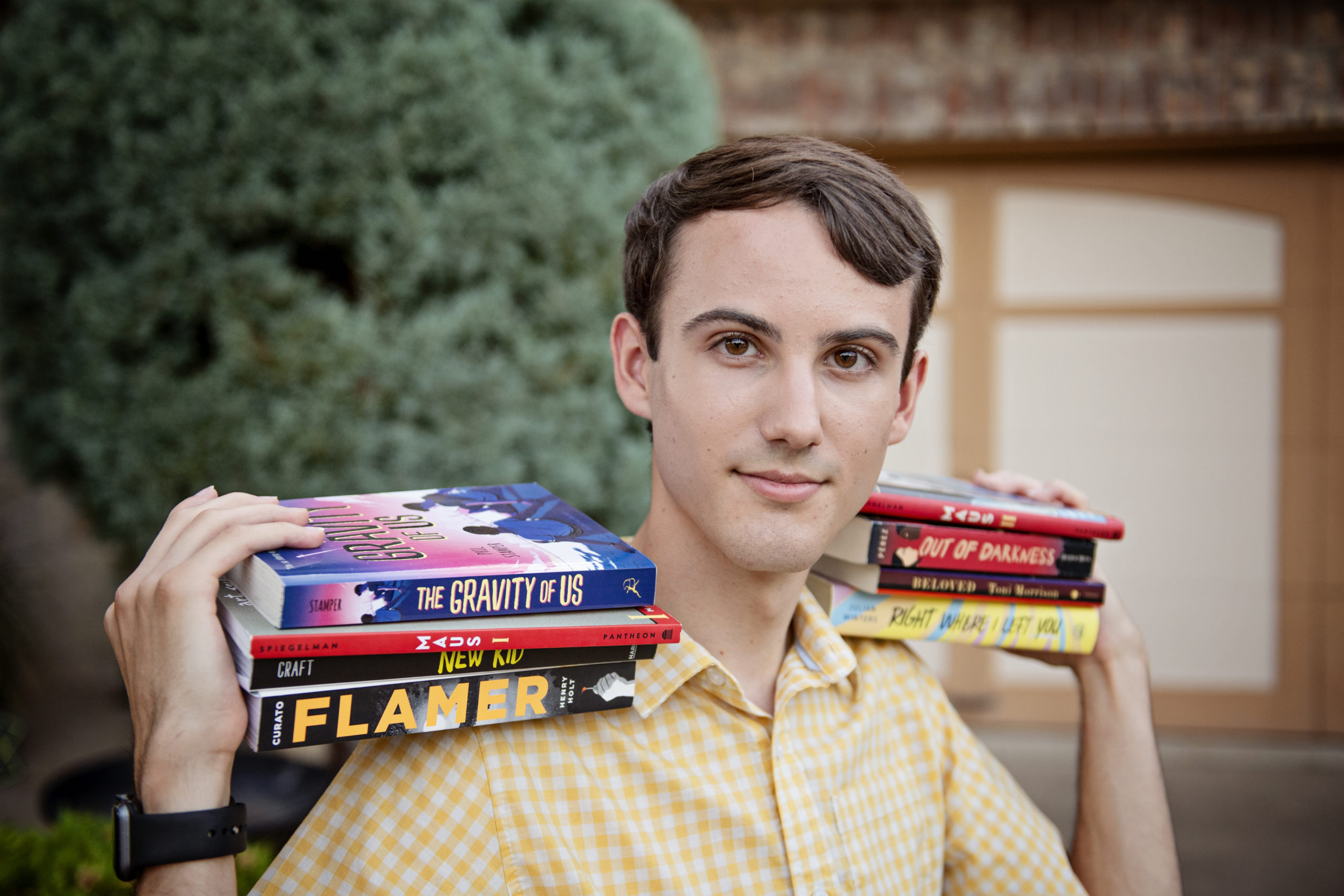 Cameron is a white person with short brown hair , wearing a button down yellow shirt, and holding two armfuls of books on their shoulders, including "The Gravity of Us," "Maus," "Flamer" and other victims of Texas book bans.