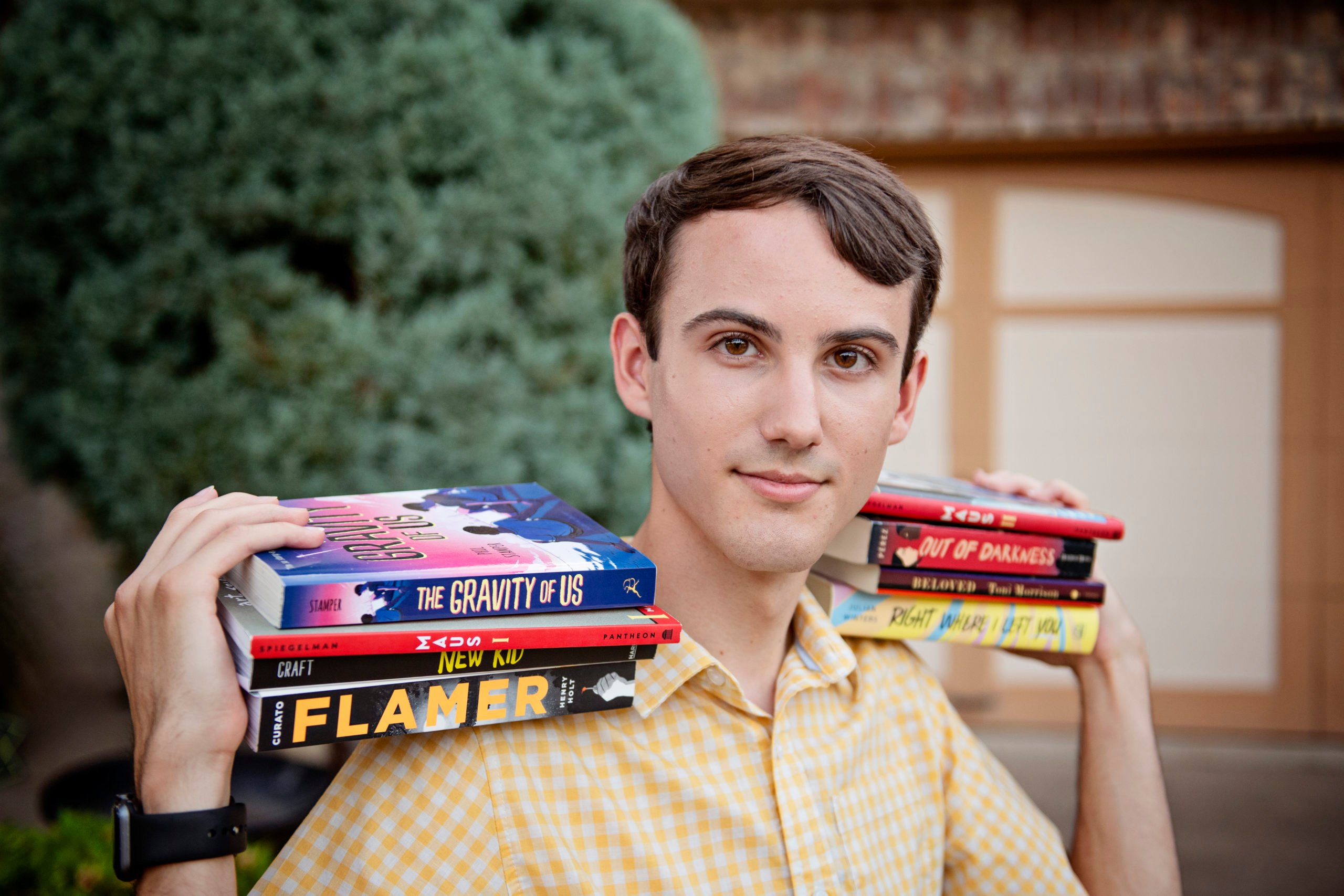 Cameron is a white person with short brown hair , wearing a button down yellow shirt, and holding two armfuls of books on their shoulders, including "The Gravity of Us," "Maus," "Flamer" and other victims of Texas book bans.