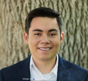 Carlos Turcios, a smiling young Latino man with short brown hair, wearing a blue suit jacket and open-collared button-down.