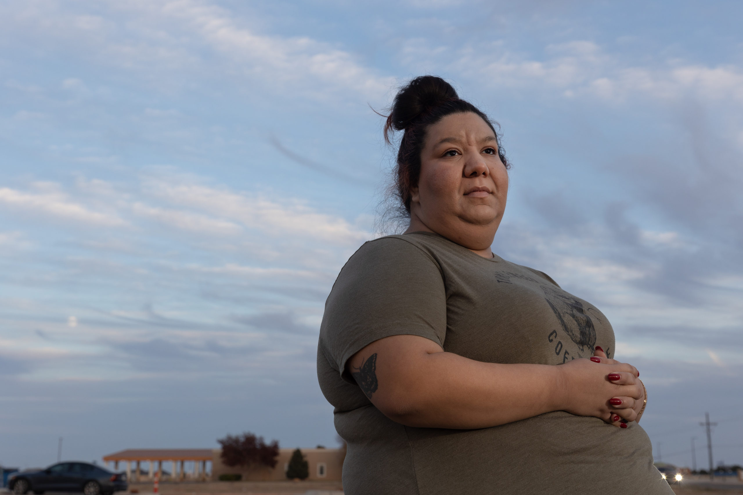 Destiny Adams stands for a portrait outside of her shop, Tumbleweed + Sage Coffeehouse, in Wolfforth, TX on December 7, 2022. She's wearing a Tumbleweed + Sage t-shirt, which shows a beaver chewing on a branch, and the sky is a speckled with moody clouds behind her.