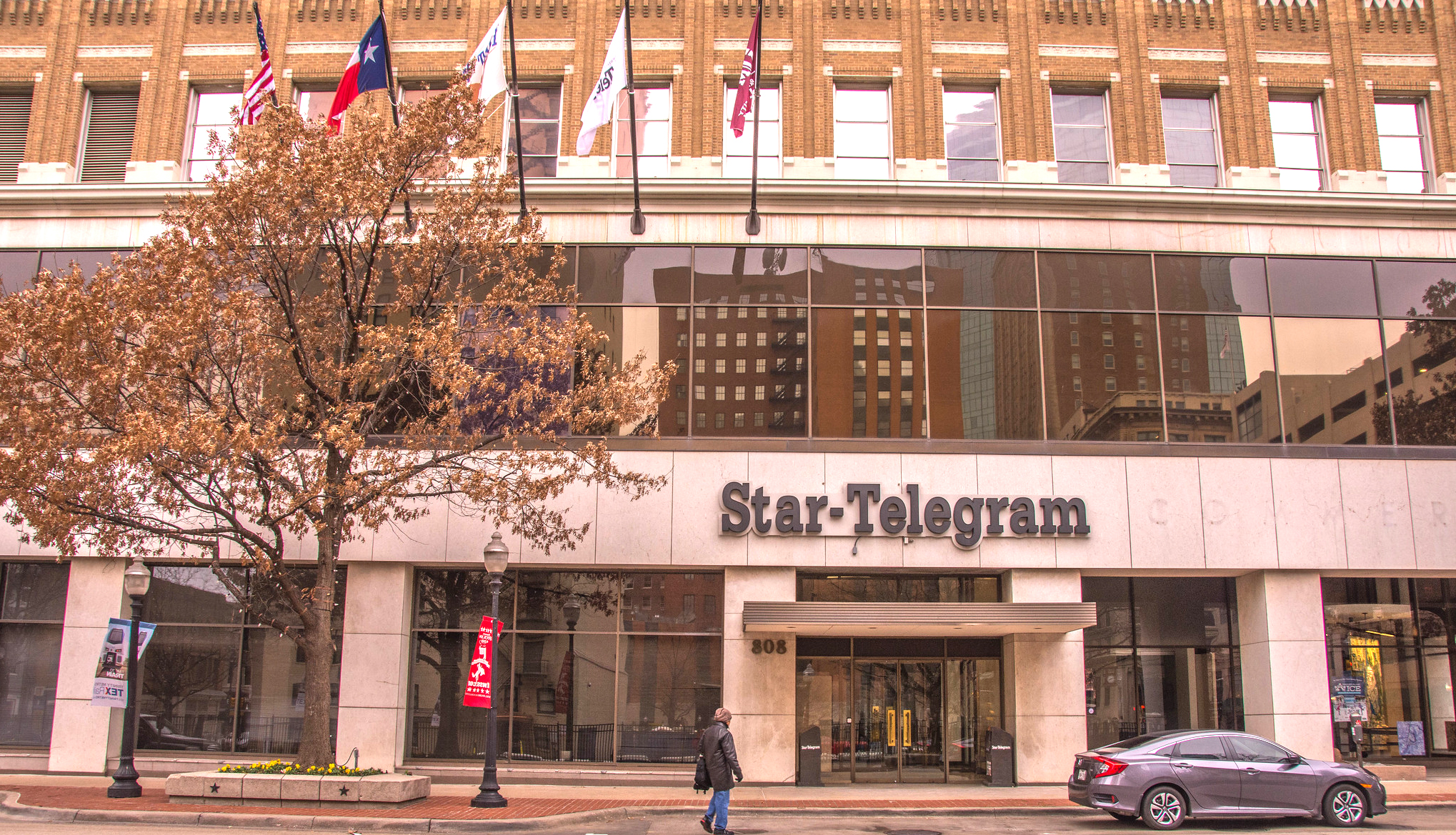 A lone person approaches the glass front doors of the Fort Worth Star-Telegram building in downtown Forth Worth, Texas. A parked car and a yellow autumnal tree stand outside.