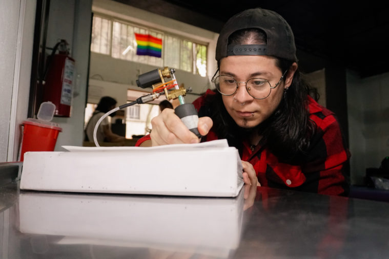 A trans man with long dark hair, in a backwards ball cap, uses a tattoo machine in a shop with a rainbow Pride flag hanging in the window. He's wearing a red and black flannel and hunched over his artwork.