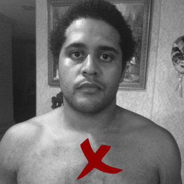 A portrait of a shirtless Black man with medium-short hair, frowning at the camera. The image is in black and white, but a colored red X marks the spot where Alan Pean got shot.