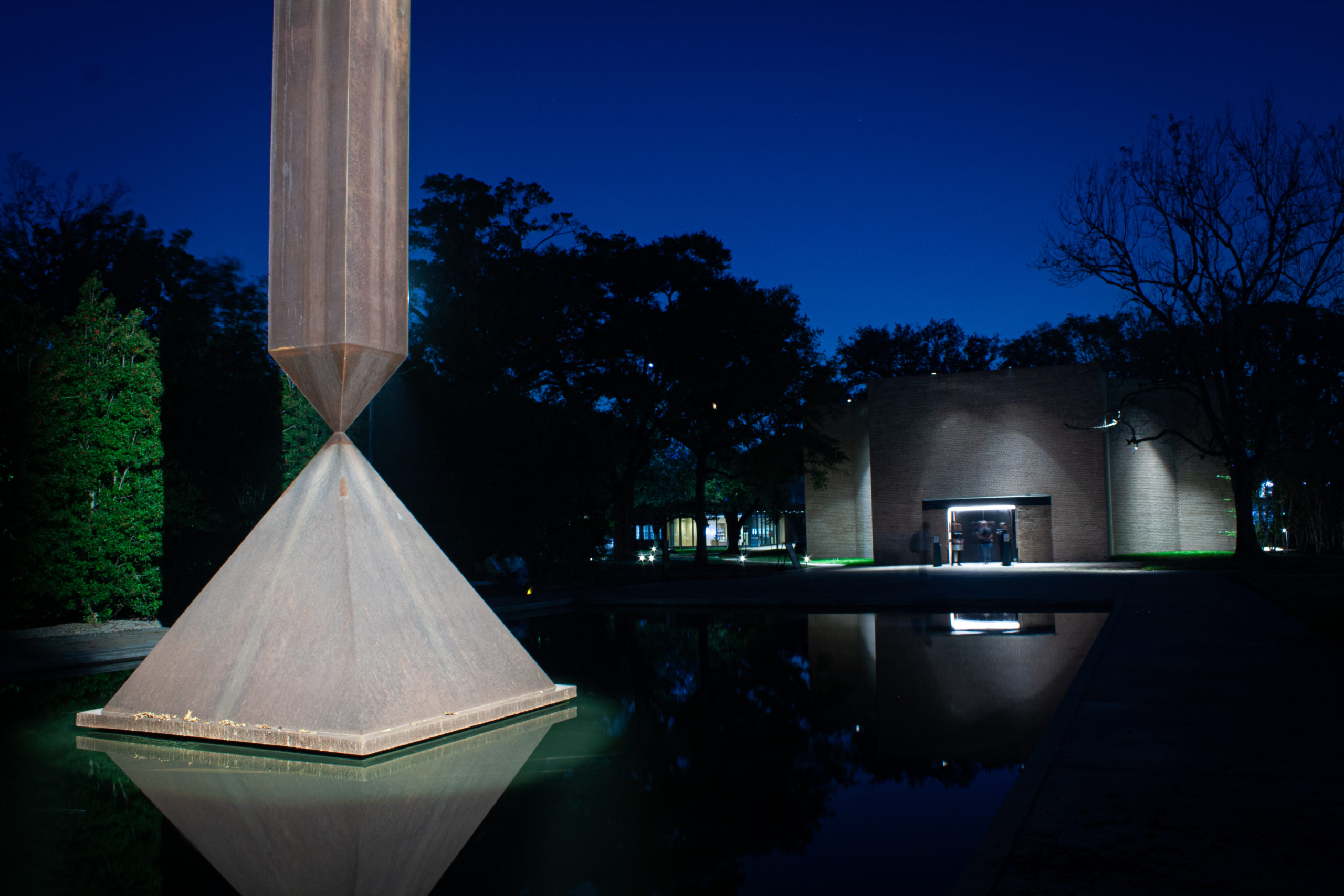 The "Broken Obelisk" sculpture and its reflecting pool stand in front of the Rothko Chapel, where a few blurry figures can be seen at the entrance. It's night time in Houston.