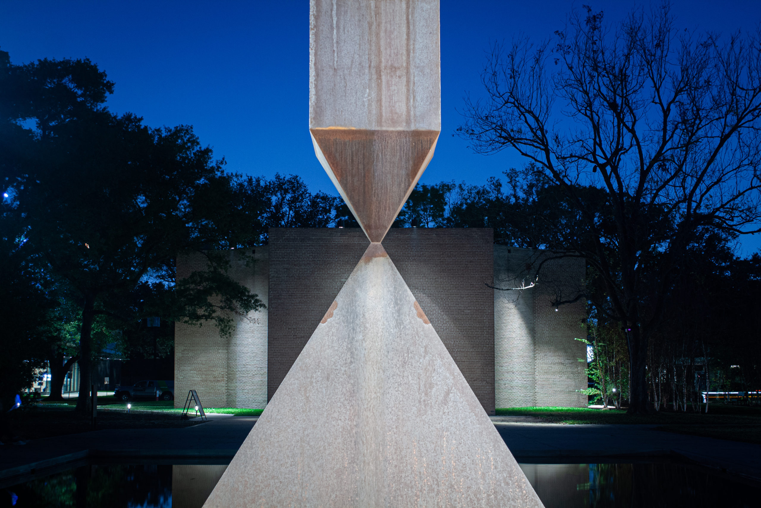 A broken granite obelisk balances, seemingly impossibly, on its point upon a pyramid of granite. This sculpture stands before Houston's Rothko Chapel, a rectangular grey brick building, seen under a clear sky at dusk.