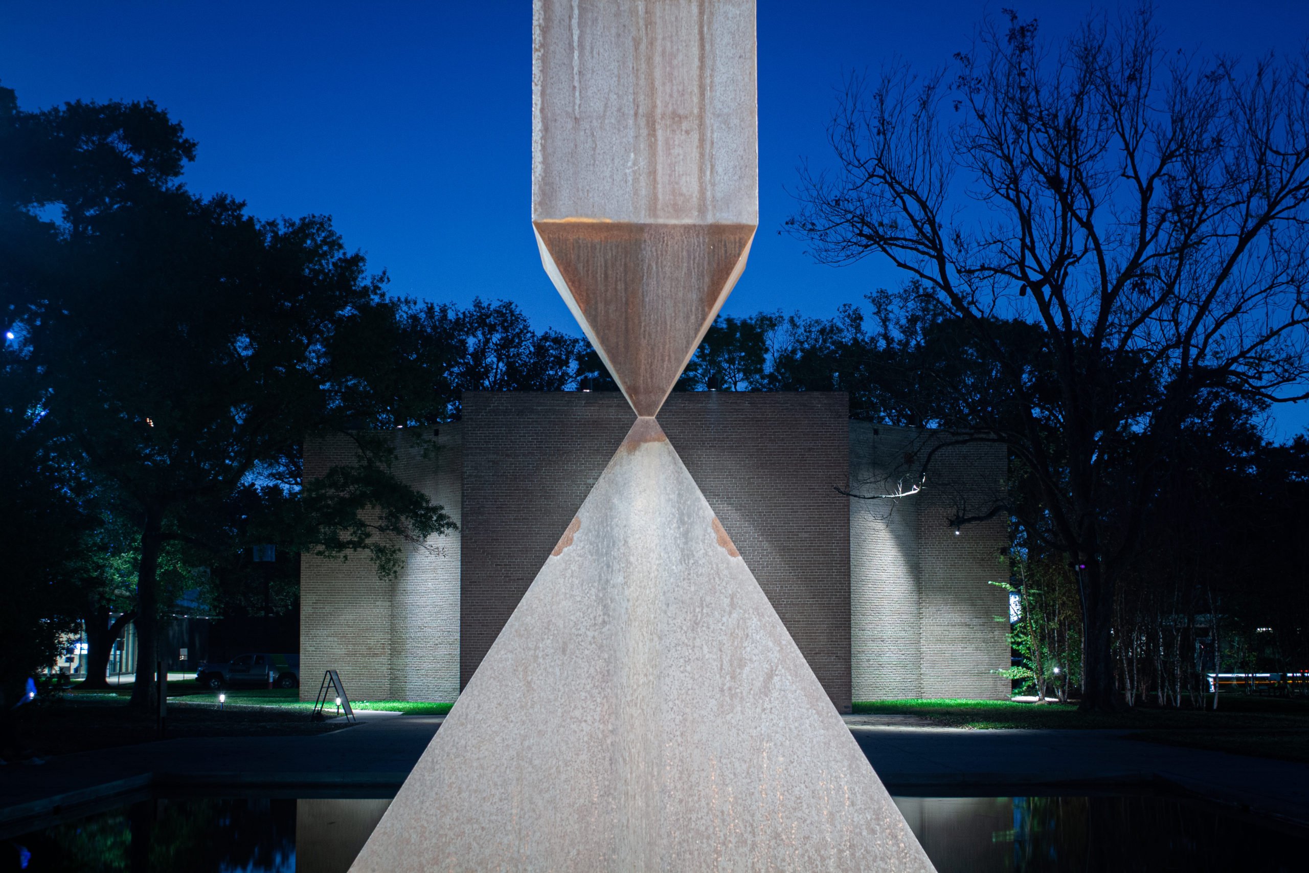 A broken granite obelisk balances, seemingly impossibly, on its point upon a pyramid of granite. This sculpture stands before Houston's Rothko Chapel, a rectangular grey brick building, seen under a clear sky at dusk.