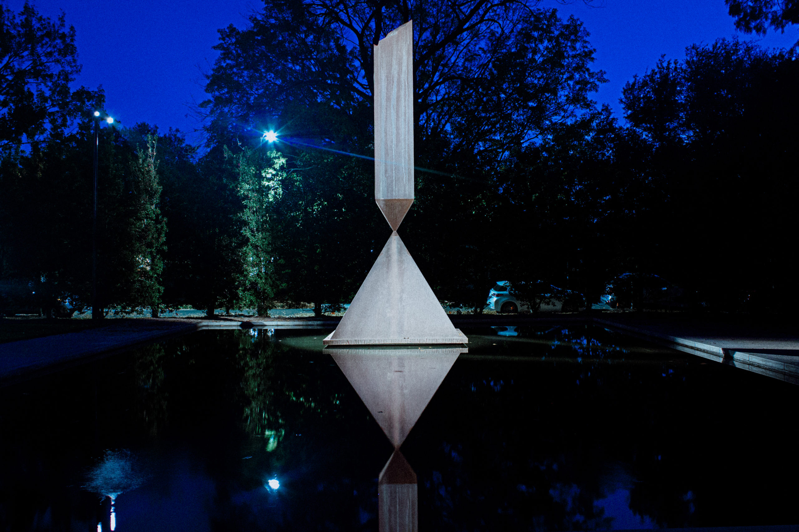 This sculpture, outside the Rothko Chapel, features a broken obelisk balancing on its point upon a pyramid of granite, placed in a lit reflecting pool surrounded by trees.