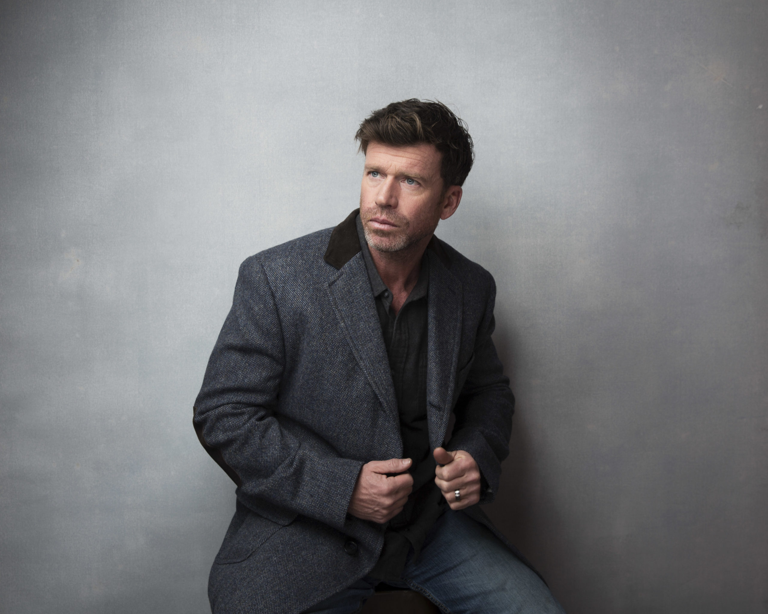 Taylor Sheridan, a white man with short brown hair in a dark suit and button down without a tie. His hands are on the lapels of his suit as he poses against a gray background.