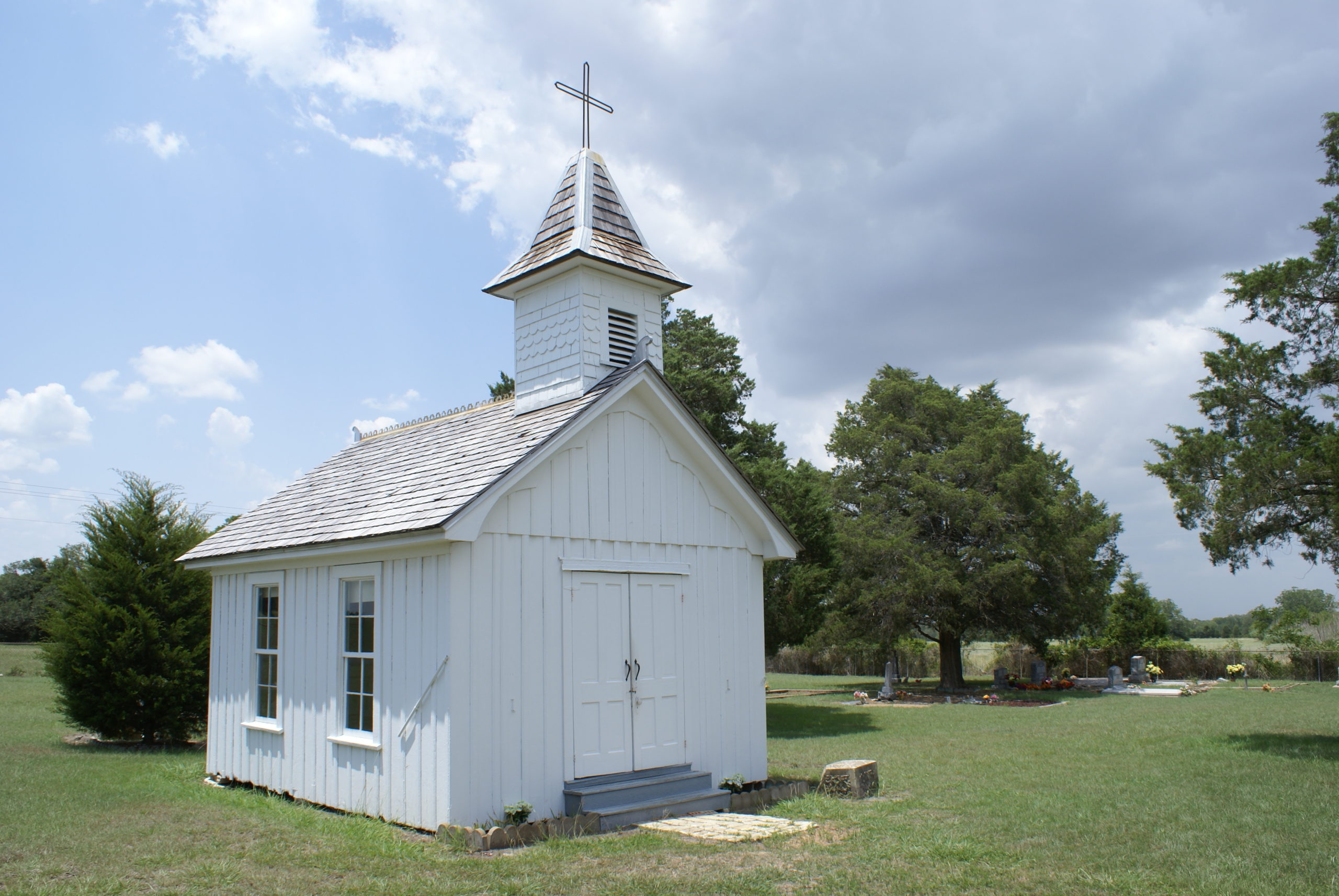 An tiny old, white-painted rustic wooden chapel stands in a Texas field under a sky with storm clouds rolling in.