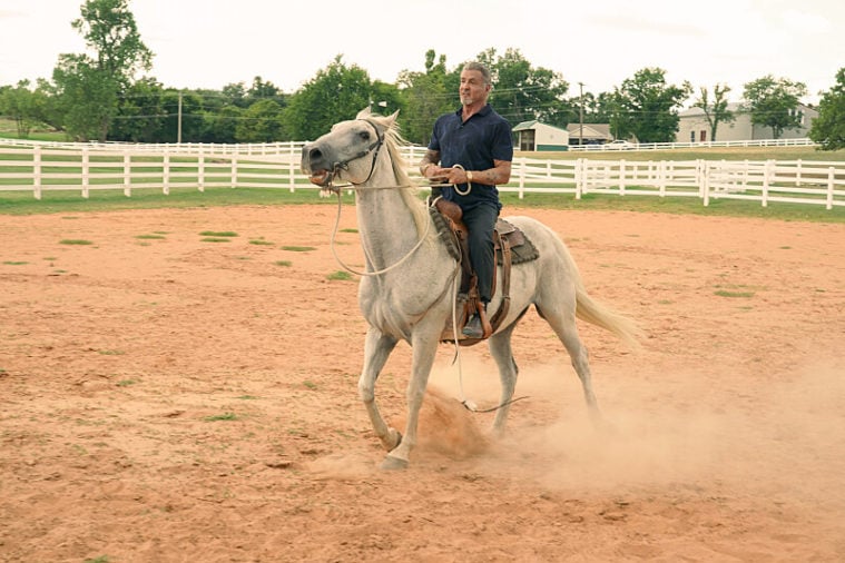 Sylvester Stallone rides a white horse across a dusty ring, wearing blue Western clothing.