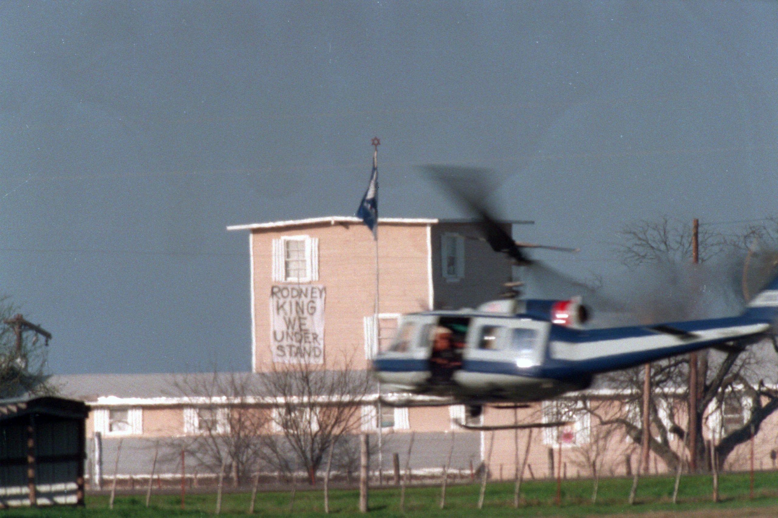 A Texas Department of Public Safety helicopter buzzes past the Mount Carmel Branch Davidian compound in this March 27, 1993, file photo taken near Waco, Texas. The sprawling compound appears to be made of stucco, with a long ranch-style area and a multi-story unit towering over the rest. Bare scrubby trees surround the property. A banner hanging from the tall building reads Rodney King We Understand.