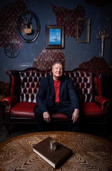 Jeff Guinn, author of the new book on the Waco siege, is a white man in a suit coat and pants, with a red shirt underneath, seen sitting on an elegant red couch in a fancy sitting room with a steampunk vibe.