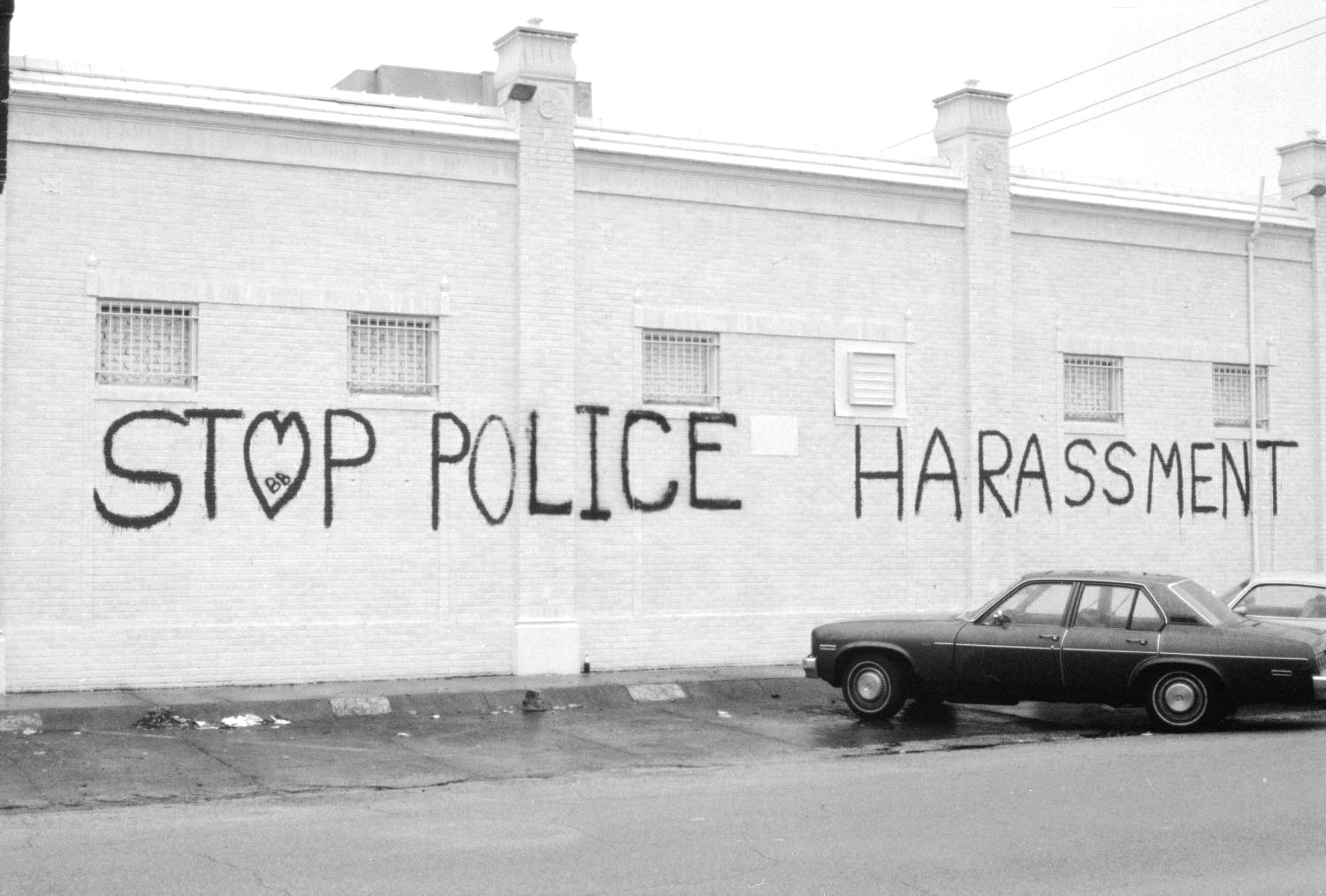 The side of a white brick building with graffiti on the side reading "Stop Police Harassment." The O in Stop is shaped like a heart. An old black car is parked near the wall. Before Lawrence, police raids and even brutality were commonplace in Texas.