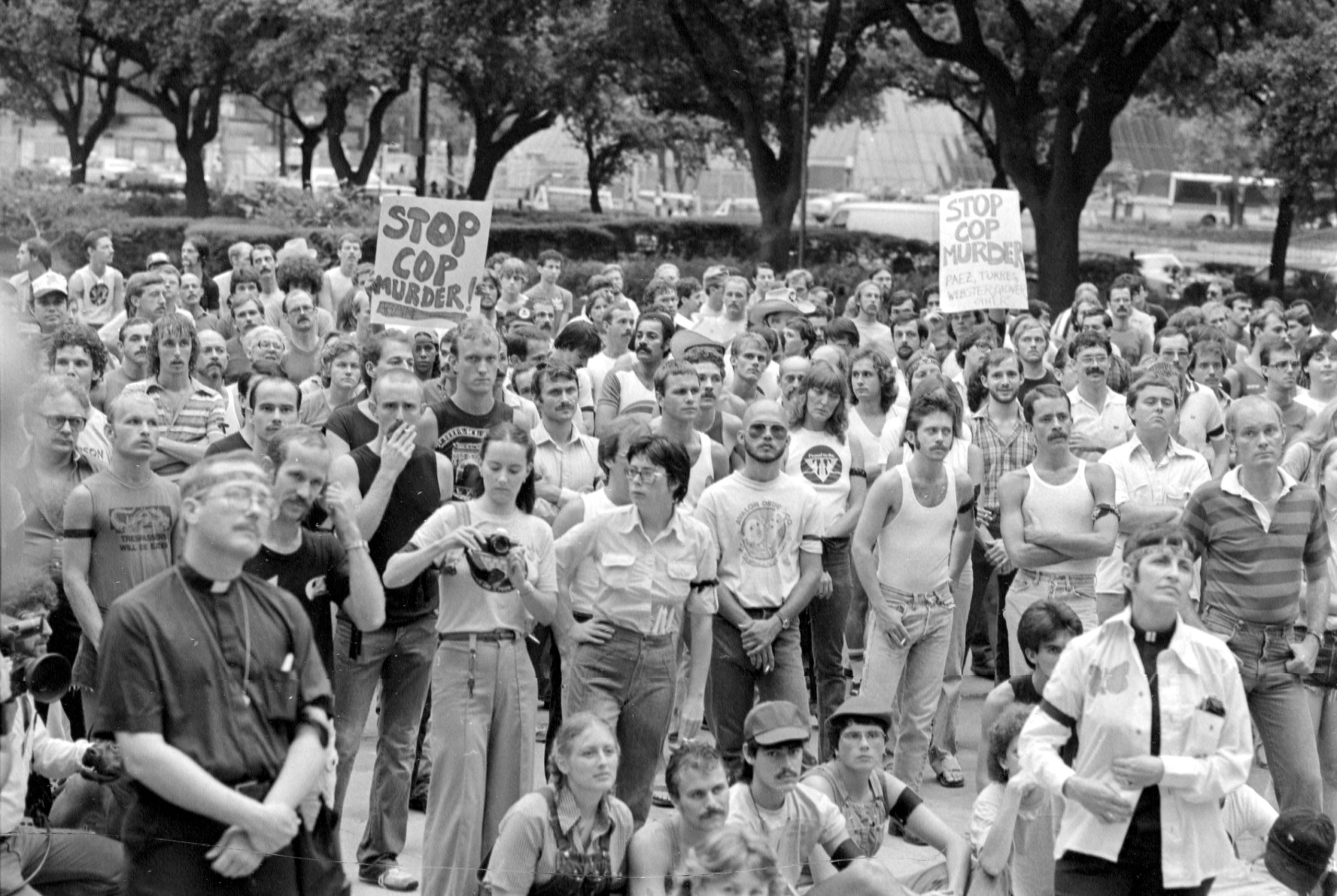 A black and white photo of a massive protest crowd in Houston, with a priest conspicuously near the front of the crowd. Signs read "Stop Cop Murder!"