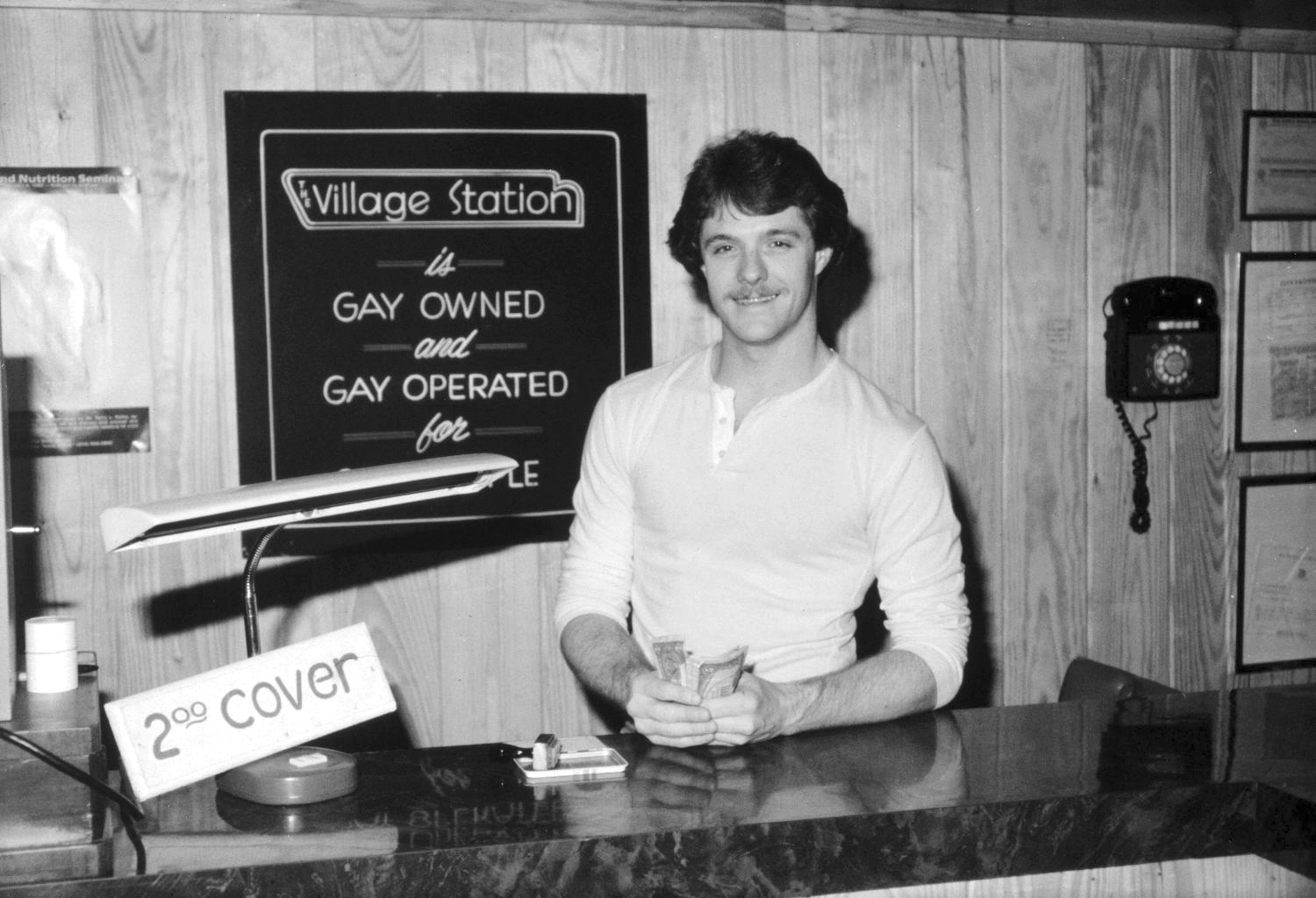 In Dallas' Village Station, a pre-Lawrence gay club, a smiling white man in a long-sleeve white shirt stands behind a counter, holding a handful of $1 bills. A sign behind him reads "Village Station is Gay Owned and Gay Operated for Gay People". Another sign proclaims that there is a $2 cover. An old fashioned lamp is on the counter and an old rotary phone on the wall.