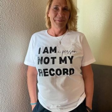 Lori Mellinger is a blonde woman in a white t-shirt reading "I am a person not my record".