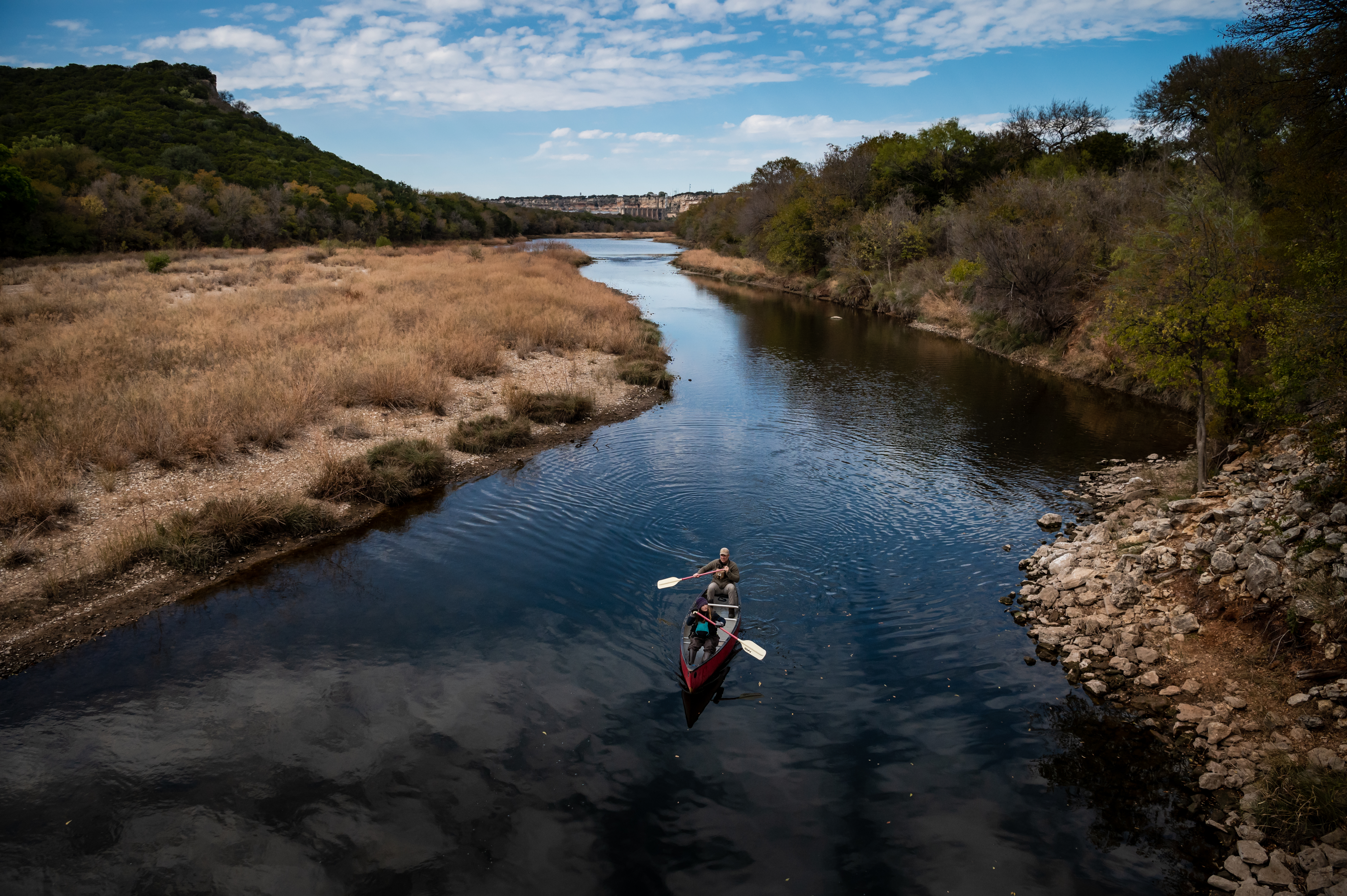 A canoe floats down the Brazos River under a big partly cloudy blue sky, with two people rowing it. The shore, covered with rocky scrub and low forested hills borders the river.