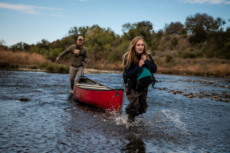 A white woman with dirty blonde hair pulls a red canoe, with the help of her father seen in the background, along a stretch of the Brazos River.