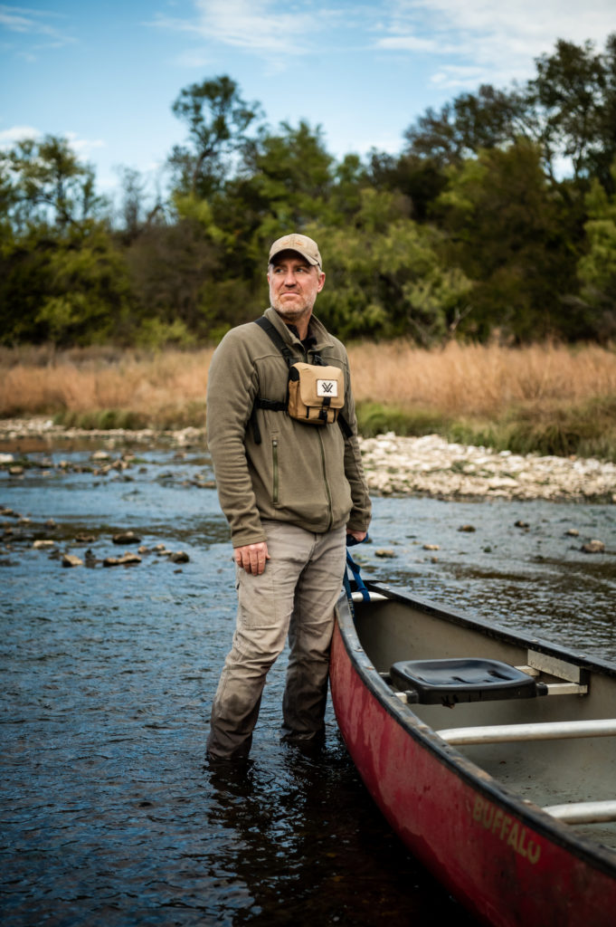 Nick Dornak, a middle-aged white man, the president of the nonprofit group Friends of the Brazos River poses for a portrait during a canoe trip down the John Graves Scenic Riverway. He's standing by a rowboat in the Brazos River, wearing a hoodie, a muddy pair of khakis and a pouch around his neck. There's a baseball cap on his head and a serious expression on his face.