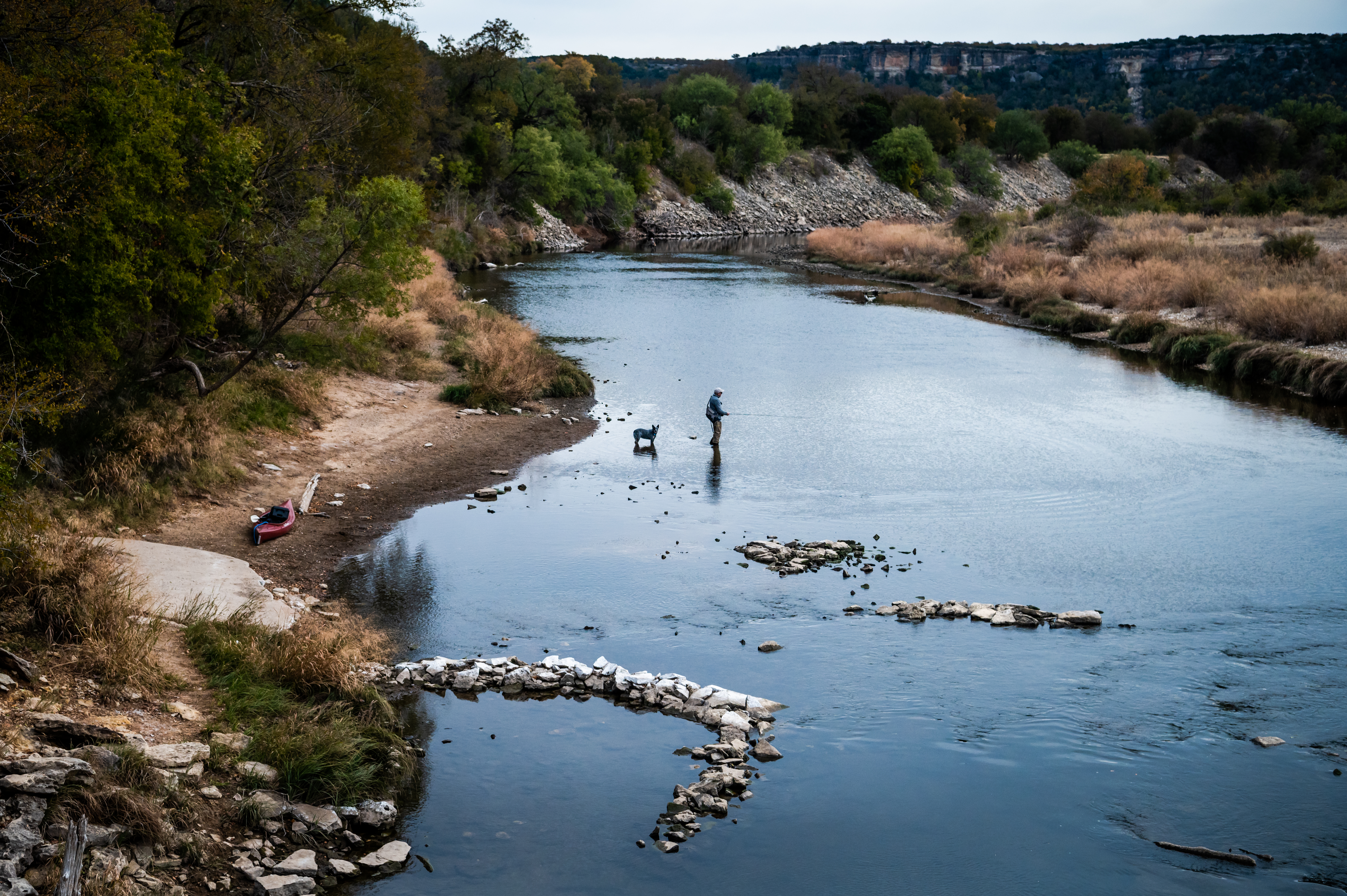 A man fly fishing with his dog in the Brazos River, surrounded by tall forested hills covered in scrubby trees. 