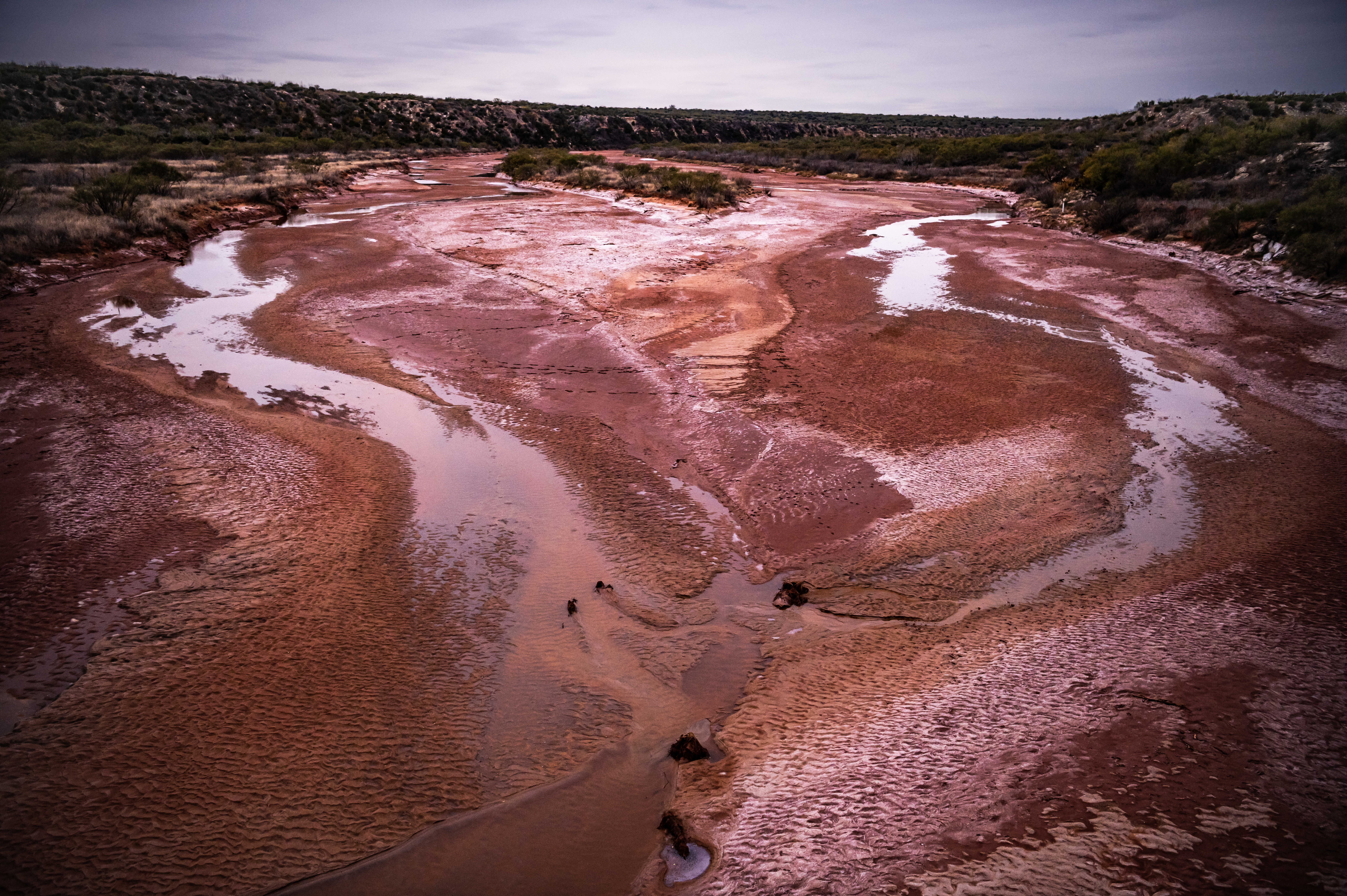 A broad expanse of waterway that is almost dried up, covered in red mud and puddles.