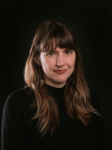 Sara Hutchinson is a white woman with brown hair just past her shoulders, posed in a dark shirt against a dark background. She's wearing long earrings and a slight smile.