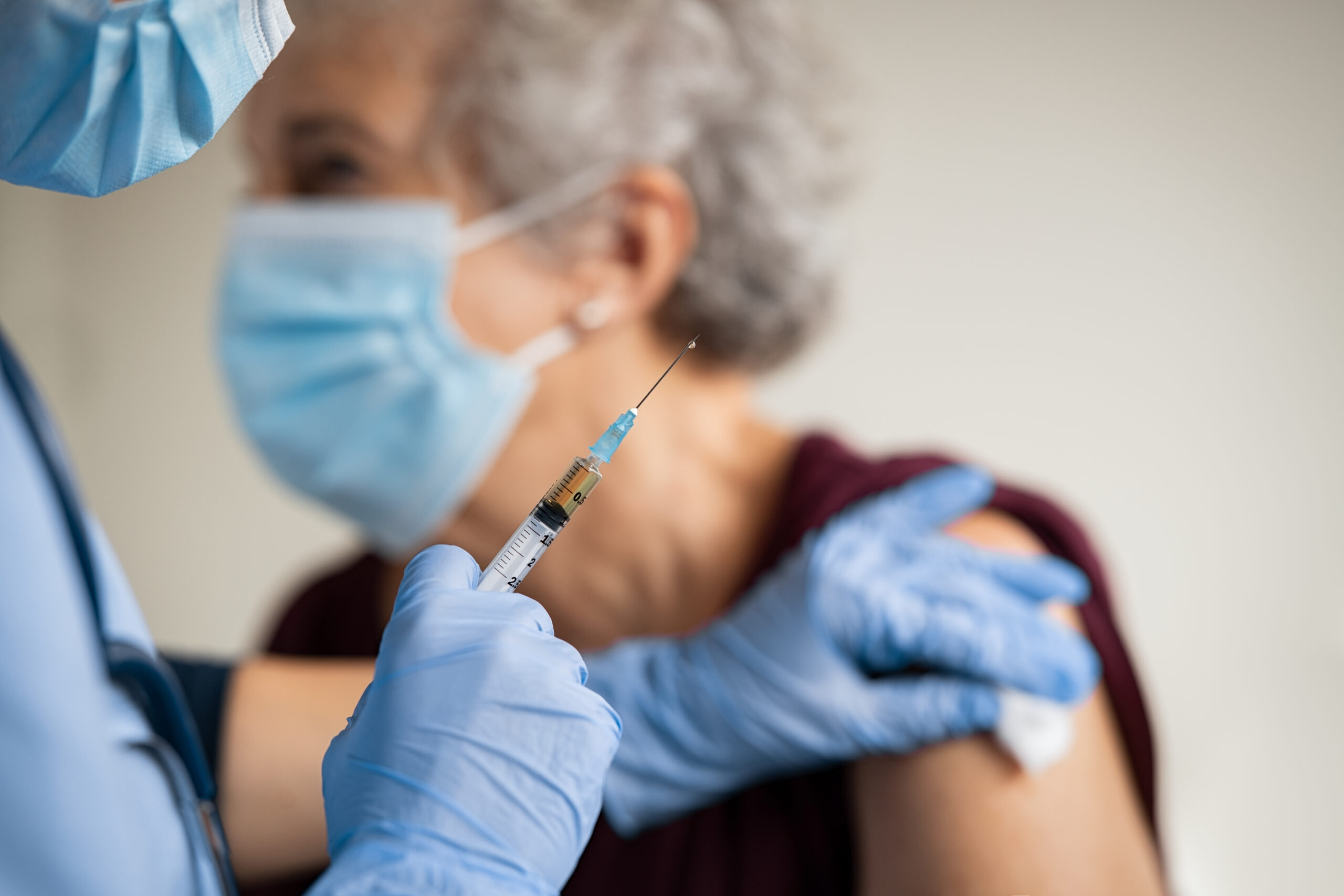 A closeup of a needle loaded with a vaccine, held in the hand of a medical professional wearing blue gloves. In the background, they are applying an alcohol swab to a grey-haired person who will receive the vaccine in their shoulder. There's increasing hesitancy where once there was widespread acceptance of vaccines in Texas.