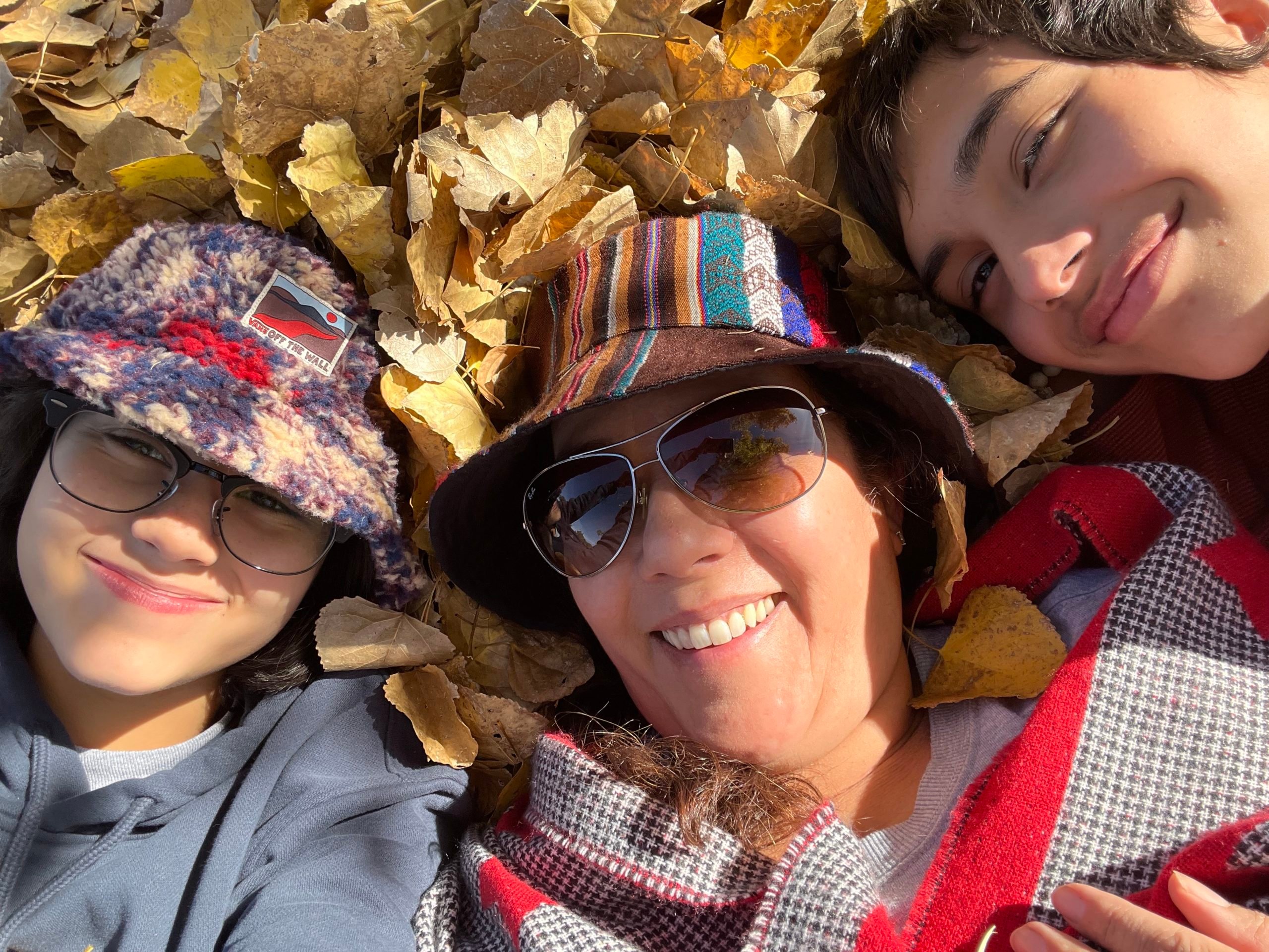 An adult woman poses in a pile of autumn leaves with two children, a boy to the right and a girl to the left, all in cozy clothing.