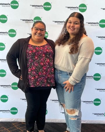 Maggie Luna, a Latinx woman, poses to the left of her daughter, who is taller than her, on a red carpet style walkway.