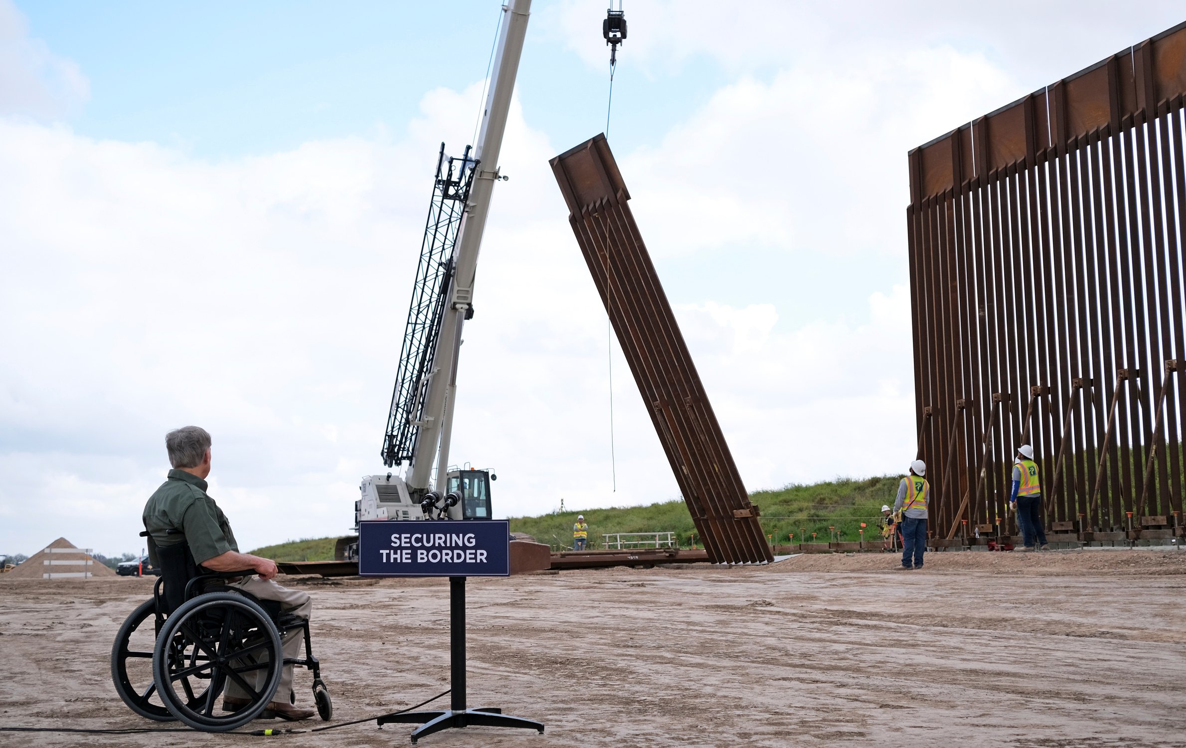Greg Abbott, sitting in his wheelchair at a podium labeled "Securing the Border," watches as a crane puts up a segment of the border wall, part of the seemingly endless spending on border security.