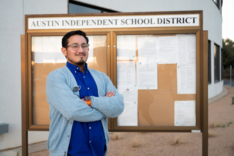 Andrew Gonzales in a blue button down sweater over a dark blue button down, and wearing horn-rimmed glases. He stands with his arms crossed and a smile on his face. He is standing in front of a Austin Independent School District bulletin board.