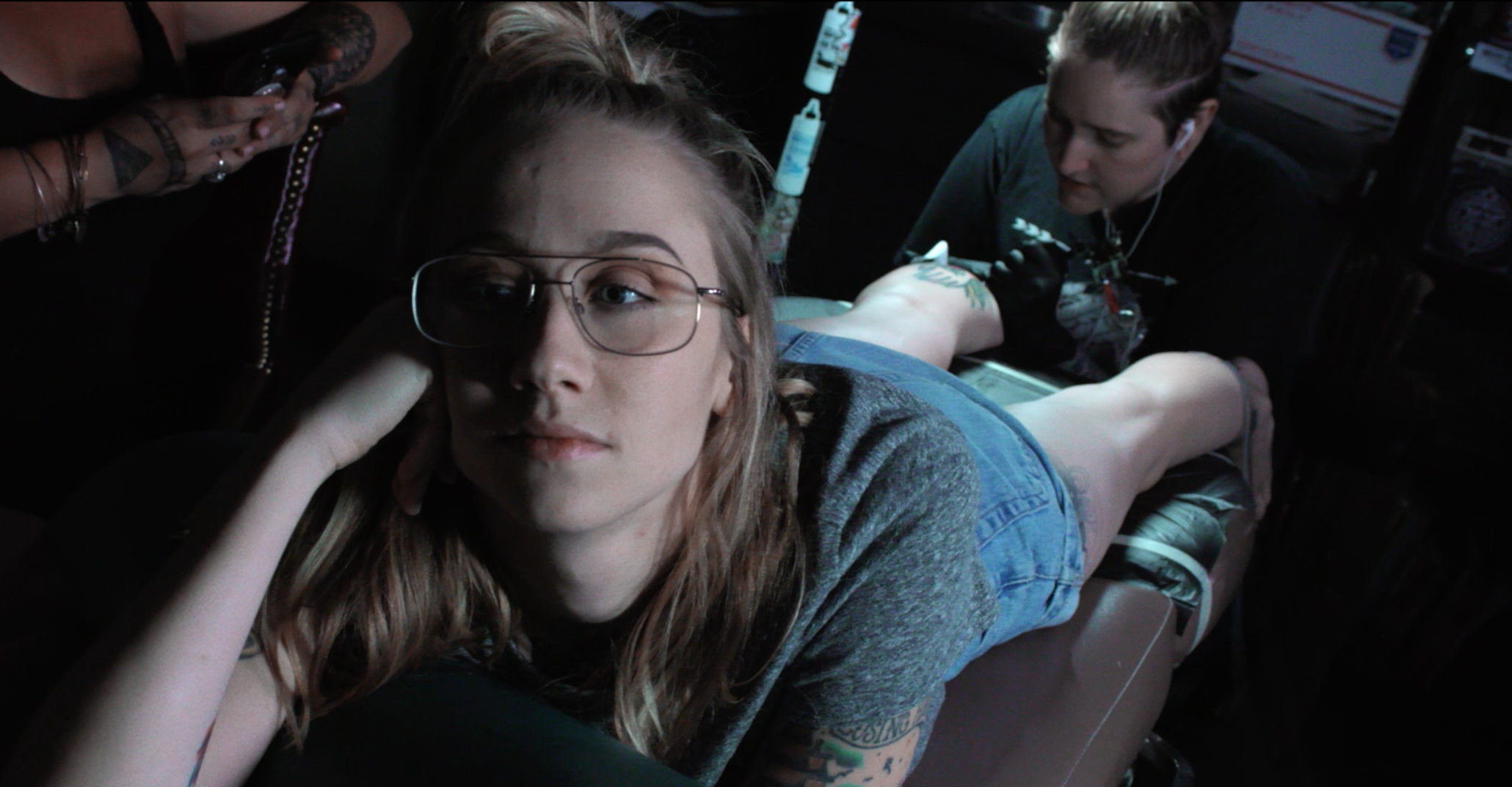 A white woman, Emily Nestor, the star of Citizen Sleuth, is seen reclining as she receives a tattoo on the back of her calf.