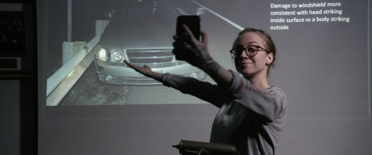 Emily Nestor, a white woman in a sweatshirt, gestures to a slide of an accident, with text reading "Damage to windshield more consistent with head striking inside surface vs a body striking outside." She's holding her phone in one hand, filming herself and the screen, and has a small smile.