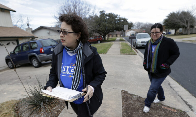 Celia Israel, a Latinx woman, is wearing a blue campaign shirt, a black and white scarf and a open black sweater. She's walking with a helper outside in a suburban neighborhood.
