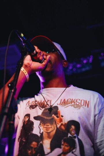 A close-up portrait of David Shabani at the microphone.  Shabani is a Black man wearing a vintage Michael Jackson tee, a black cross necklace, a baseball cap and shades.