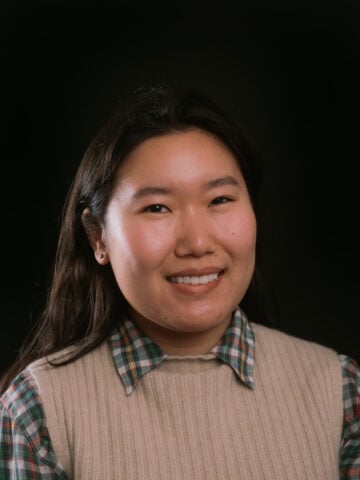 Delger is a smiling woman with black hair, wearing a light sweater vest over a checkered black, white and brown flannel.