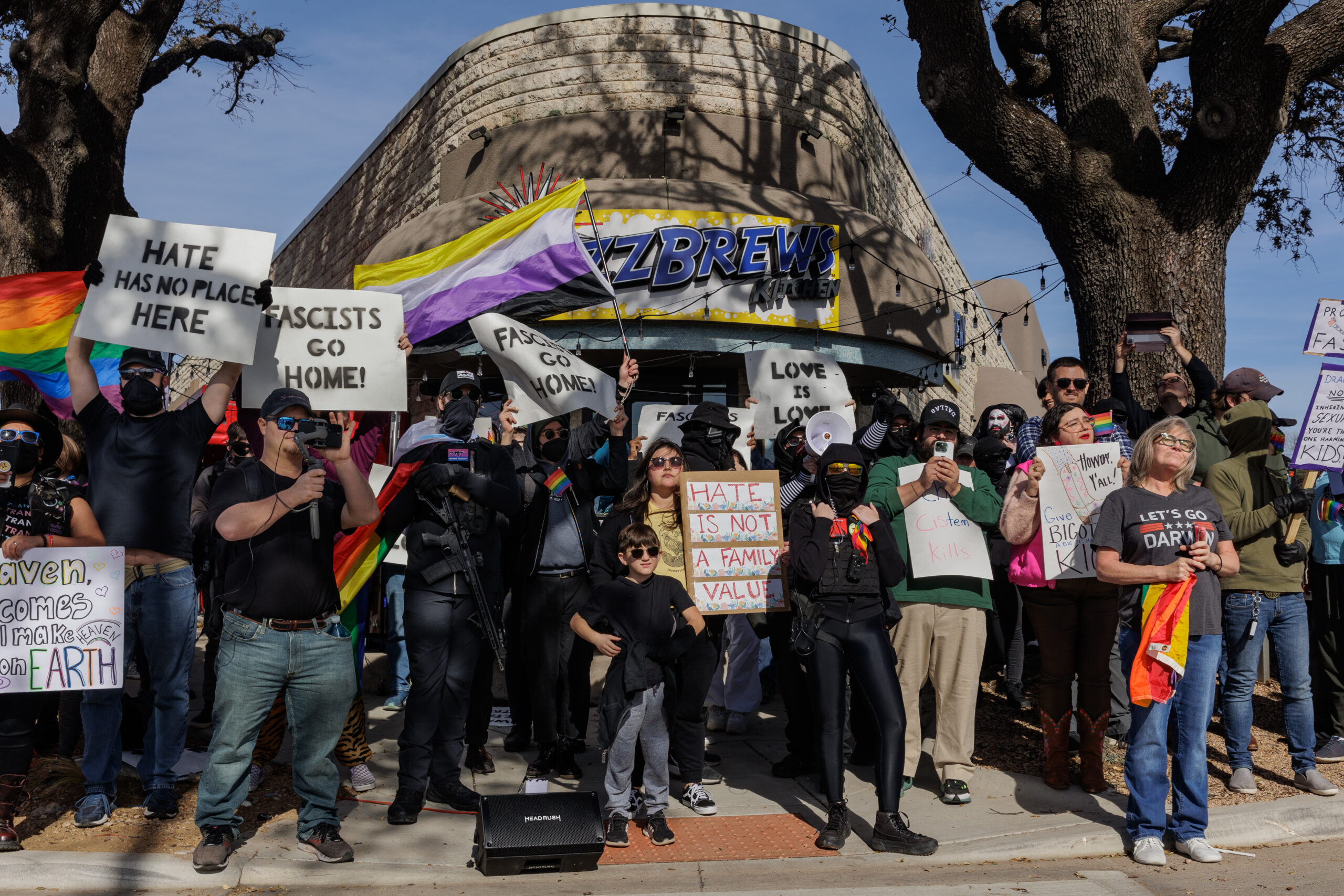 An array of community members outside "Buzzbrews" where they are protecting a drag brunch event. Some are open carrying while others have signs with mottos like "Hate has no place here" and "Fascists go home!"