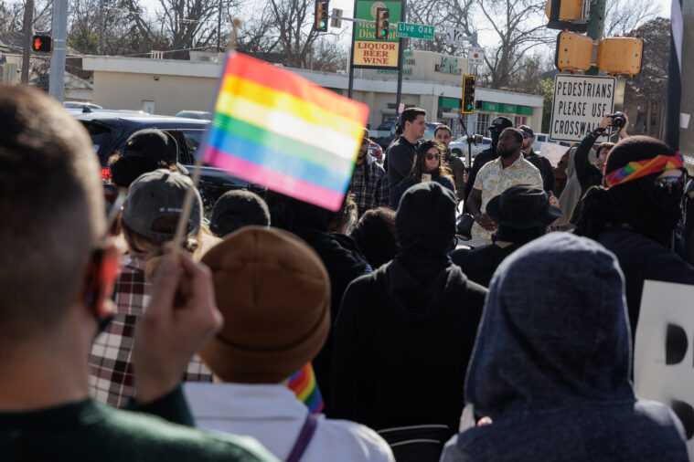 A crowd seen from behind confronts Kelly Neidert and her smaller group of supporters, including Christian youth from the New Colombia Movement. She is holding a phone and wearing a blue sweatshirt. One person confronting her is holding a rainbow Pride flag.