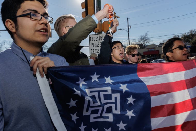 Protesters hold a modified American flag with the logo of the New Colombia Movement on it, a crusader style cross surrounded by stars. One holds aloft a rosary in his hand while others chant and shout.