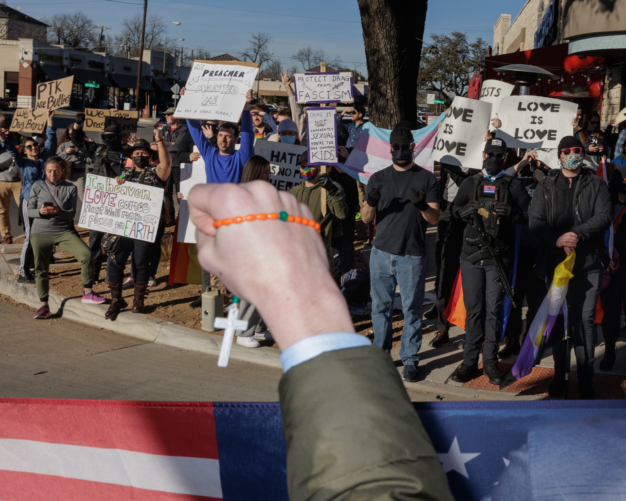 A Christian youth protester confronts supporters of LGBTQ+ people outside a "drag brunch" in the Dallas area. Seen from behind, we just see his hand in a suit jacket holding a rosary, as he stands behind an American flag.