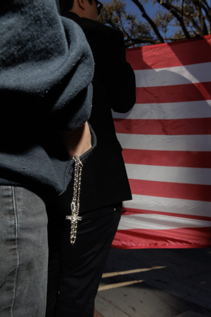 A member of the New Colombia Movement, a fascist Christian youth movement, hold a custom American flag with their logo on it. We see one member in all black, holding a rosary in his hand which is partly in the pocket of a hoodie.