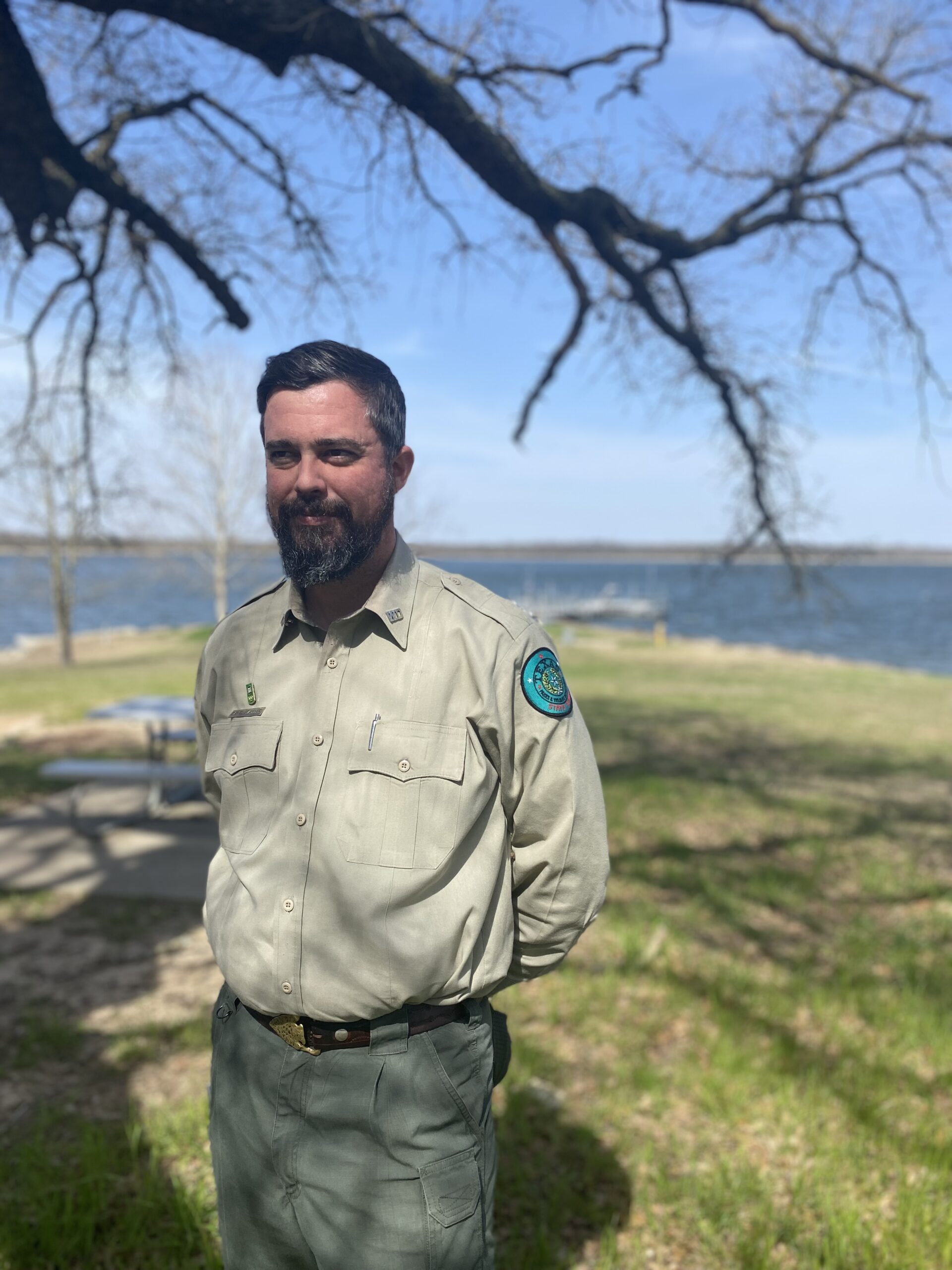 A bearded white man stands in a park ranger's uniform, his hands behind his back, with the wide open green and blue of Fairfield Lake State Park behind him. He is shaded by the branches of a leafless winter tree.