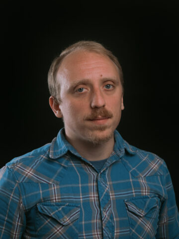 Gus Bova is a white man with short brown hair, a trimmed mustache, wearing a blue and white button down flannel.