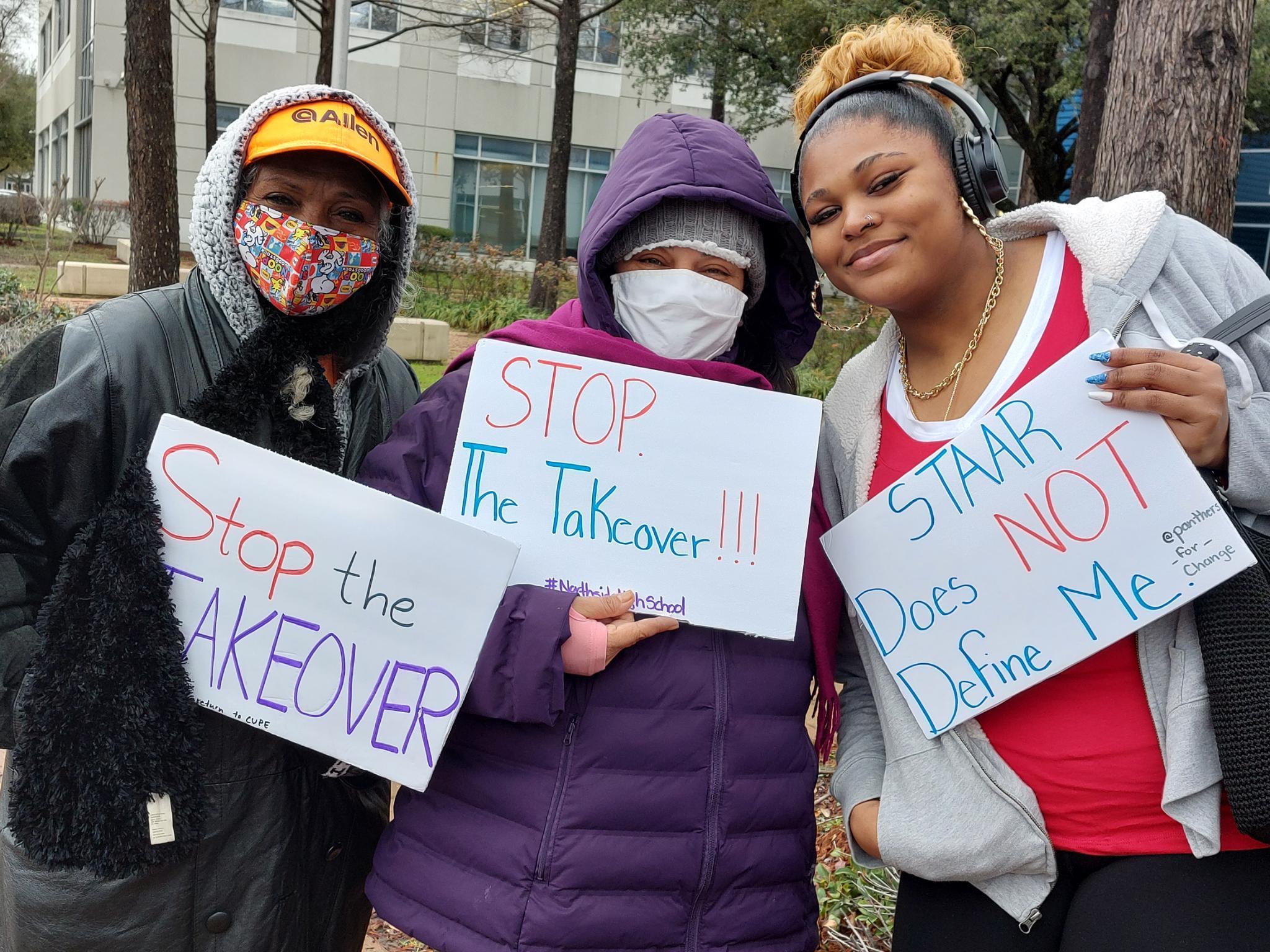 Three people gather at a winter-time protest, wearing cold-weather clothes, to protest the Houston ISD takeover. Two hold signs that read "Stop the Takeover" while another is holding a sign that says "Starr Does NOT Define Me".