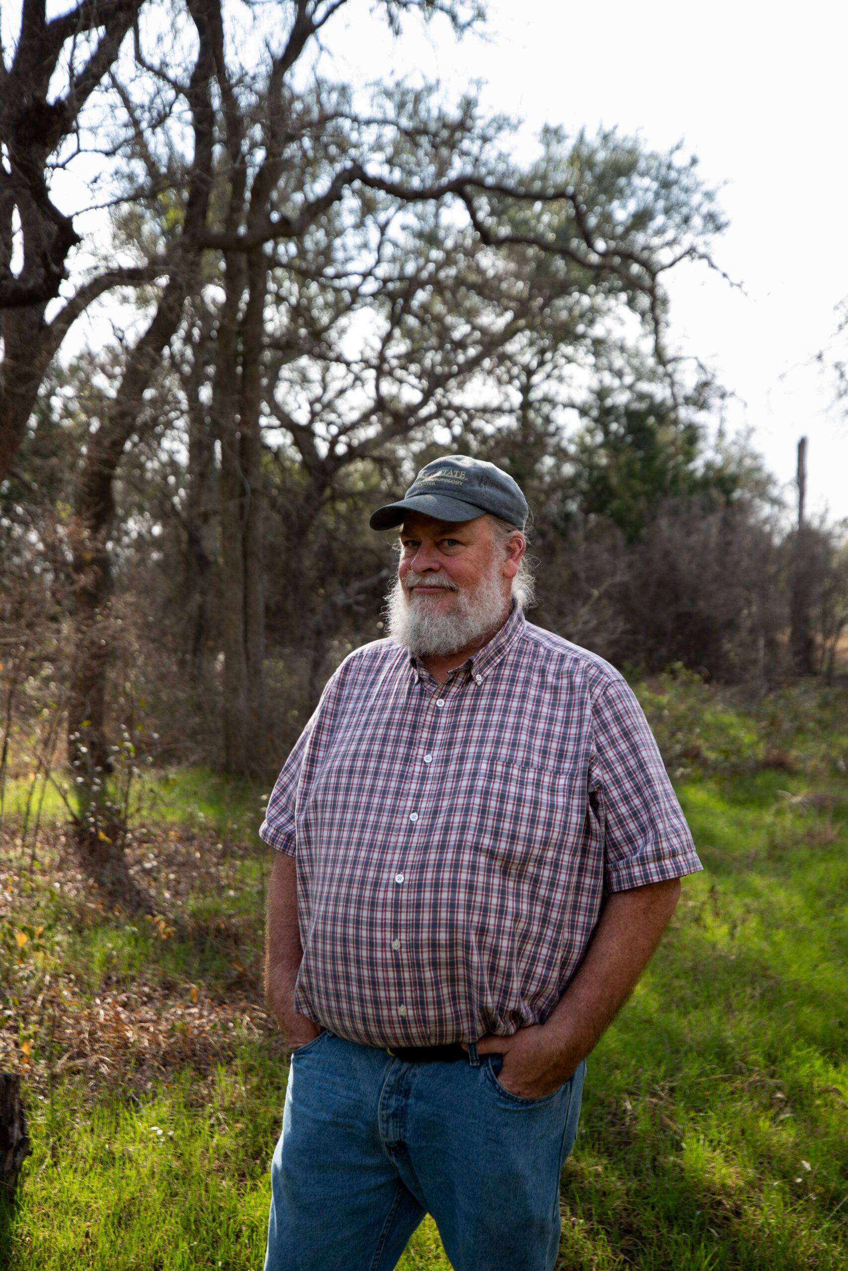 Bare oak trees crowd a small meadow where Forensic Anthropology Center Director Daniel Wescott says he would like to be placed on the body ranch as part of a study after he dies. Westcott is an older white man with a silver beard, wearing a baseball cap, jeans and a red and white flannel button-down.
