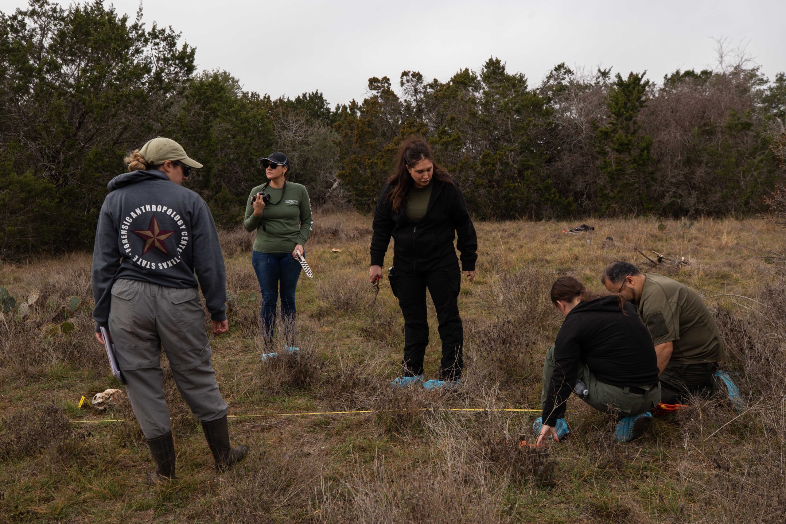 Five students gather around a group of scattered, bony human remains barely visible in the tall grass in a clearing. Two of them crouch as they take a measurement.