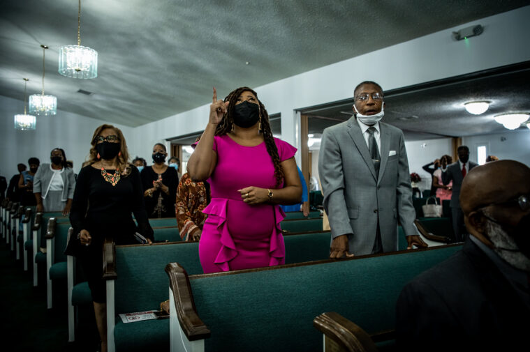 Black parishioners stand in a church as they sing hymns. Most are wearing masks. They are in rows behind gray pews under hanging glass chandeliers.