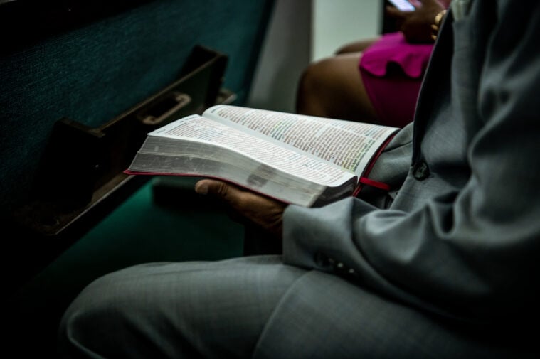 A photo of a Black man holding a bible in his lap. He's dressed in a formal suit, and sitting in a pew with other parishoners.