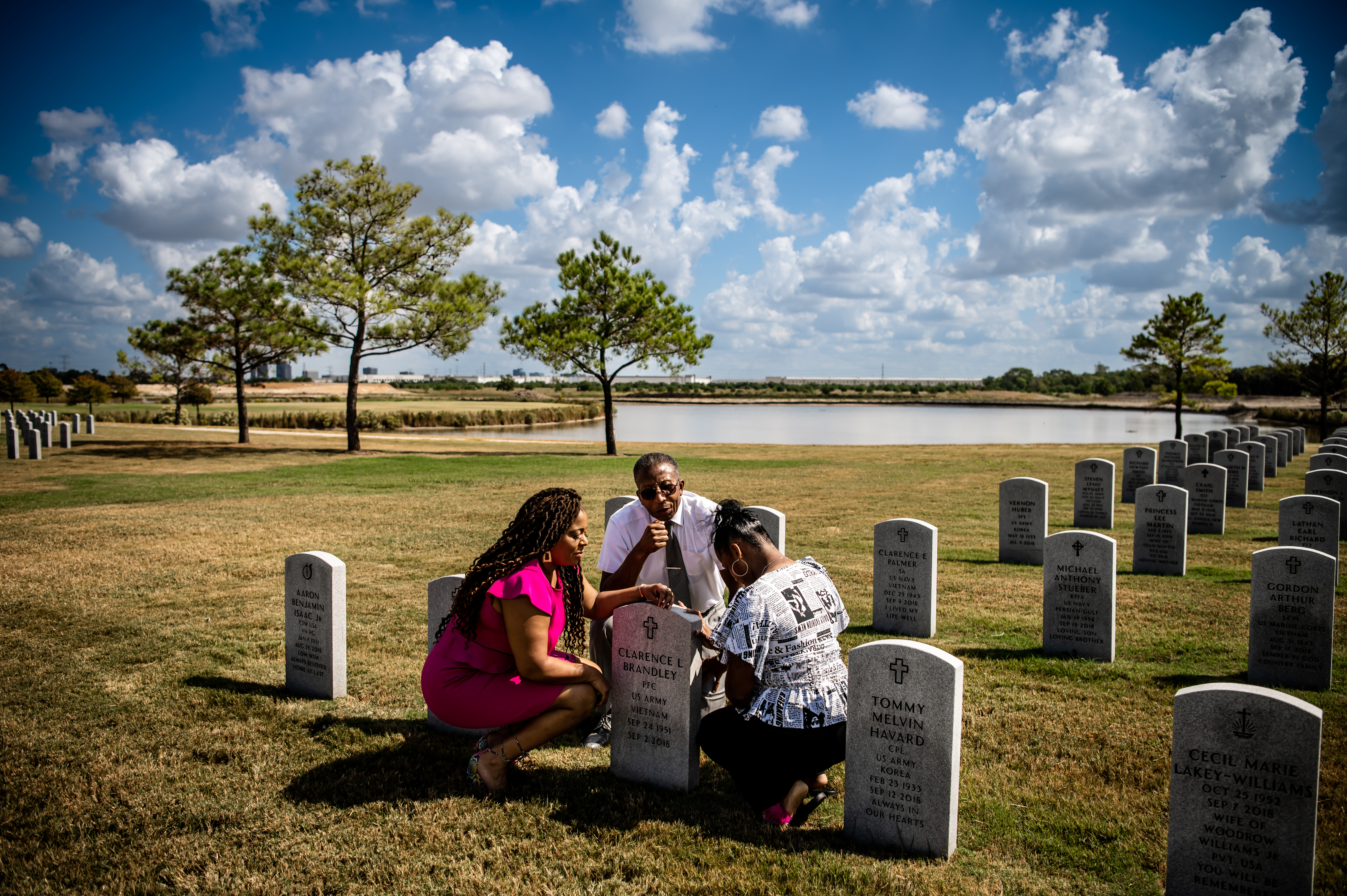 A Black family, Ozell Brandley and his nieces, Cassie Brandley and Elizabeth Phillips, pray together at the grave of his brother and the women’s father, Clarence Brandley. The graves extend away in orderly rows on a clipped lawn under a big partly cloudy Texas sky.